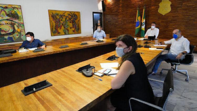 prefeito, governador, secretária de saúde e secretário sentados em grande mesa, todos de máscara